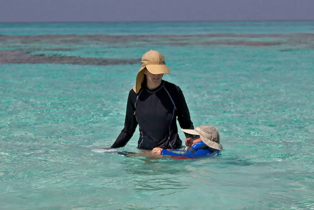 A woman standing waist-deep in clear turquoise shallows, walking a small child along on a float