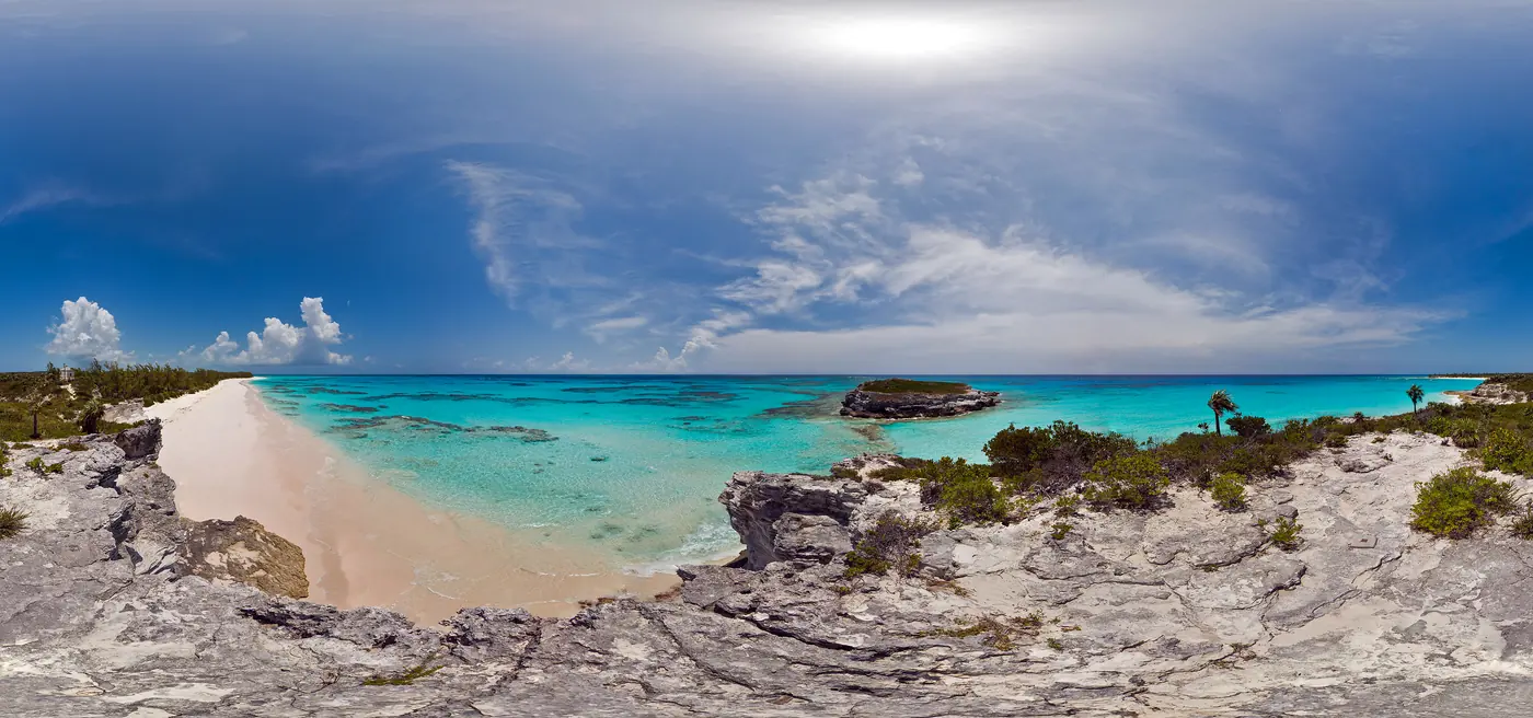 A wide view of Lighthouse Point: pink sand curving away between limestone bluffs, turquoise water broken by dark reef, and a low rocky headland out in the bay