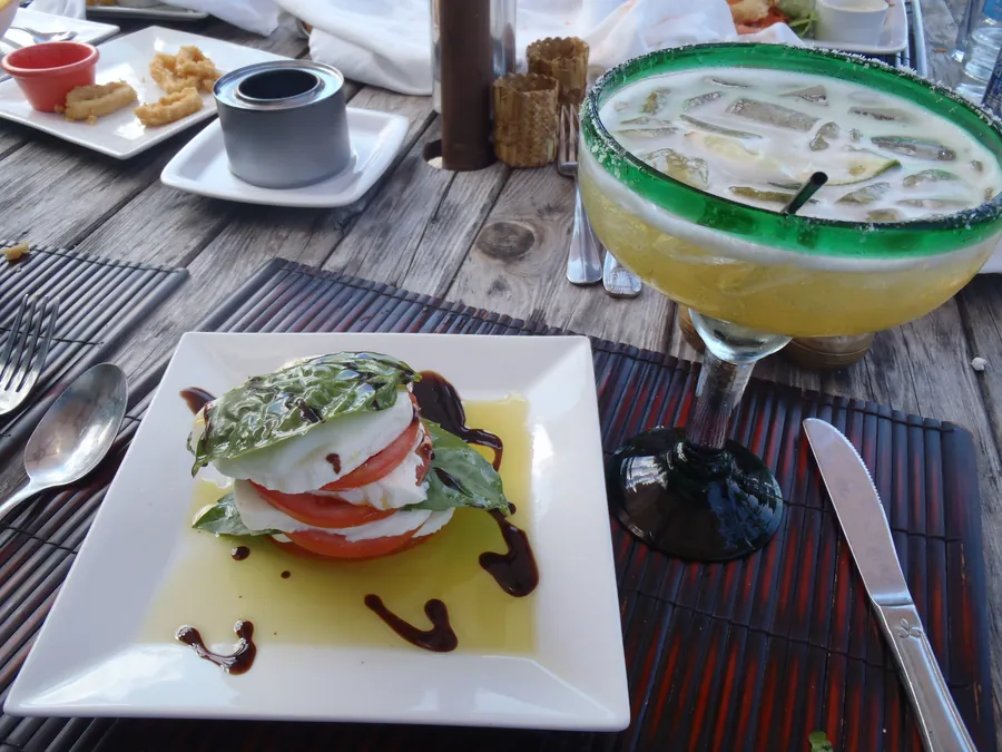 A caprese stack of tomato, mozzarella and basil with olive oil and balsamic, beside a margarita, on a weathered wooden table outdoors