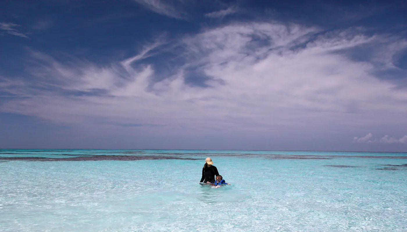 A wide sky over shallow turquoise water, a child being walked out over the sand flats