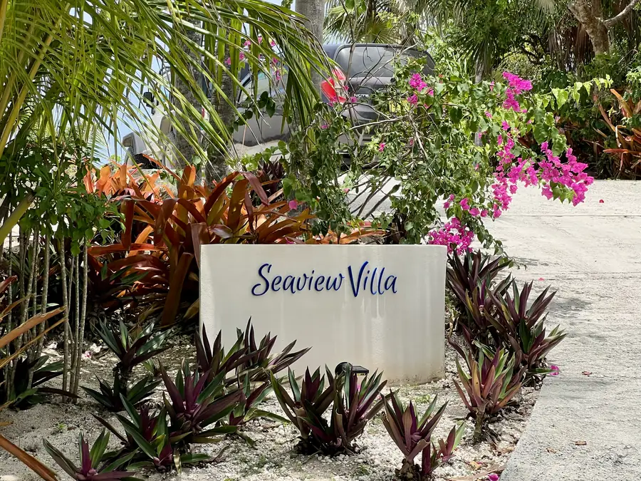 The Seaview Villa sign, painted in blue script on a white wall, in a bed of bromeliads with bougainvillea behind